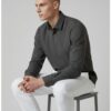 Man seated on a gray stool in a dark gray long-sleeve shirt and white pants, facing left in profile.