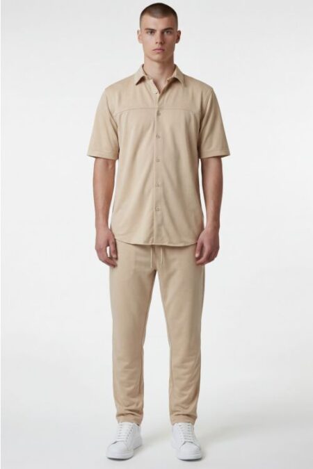 Full-body shot of a man wearing a beige short-sleeve button-down shirt and matching pants with white sneakers, standing against a white background.