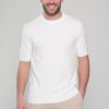 Smiling man wearing a white short-sleeve knit sweater and beige pants, standing against a light gray background.