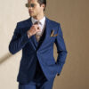 Man in a navy blue suit adjusts his tie while wearing sunglasses and a pocket square against a beige wall.