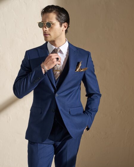 Man in a blue suit adjusting his tie while wearing round gold sunglasses against a beige wall.