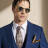 Man in a blue suit with a patterned tie and gold sunglasses, standing against a beige wall with a pocket square visible.