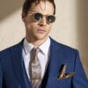 Man in a blue suit with white shirt, gold patterned tie, and pocket square, wearing round sunglasses against a beige wall