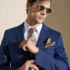 Man in a blue suit adjusts his cuff, wearing sunglasses, a patterned tie, white shirt, and a tan pocket square.