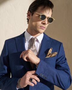 SS26-8000-BLU_2jpg - Noble Man in a blue suit adjusts a tan patterned tie while wearing round gold-rimmed sunglasses on a light background; pocket square visible.