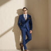 Man in a sharp blue suit with brown loafers leans against a light wall, wearing sunglasses in a sunny studio setting.