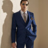 Man in a navy suit with brown tie and pocket square leaning against a light wall, wearing sunglasses.