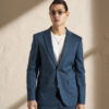 Man in a teal-blue suit with a white T-shirt, wearing sunglasses, standing against a light wall in a studio.