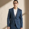 Young man in a blue suit with a white T‑shirt, round amber sunglasses, and a gold necklace, posing against a beige wall with diagonal shadow.
