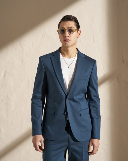 Young man in a blue suit with a white T‑shirt, round amber sunglasses, and a gold necklace, posing against a beige wall with diagonal shadow.