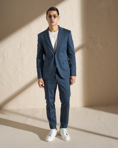 Full-body shot of a man in a blue suit with a white T-shirt, white sneakers, and sunglasses standing against a beige wall with diagonal shadow.
