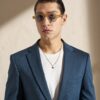 Portrait of a person wearing a blue blazer over a white t-shirt, with gold chain and round amber sunglasses, standing against a beige wall.