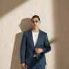 Man wearing a blue suit and white shirt, sunglasses, standing against a textured beige wall.
