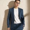 Man in a blue suit with a white tee, gold necklace, and round yellow sunglasses leaning against a beige wall.