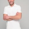 Smiling man with dark hair in a white polo shirt and beige pants crosses his arms against a light gray background.
