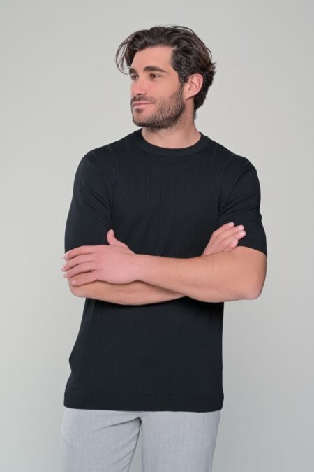Casual portrait: man with dark hair and light stubble, wearing a black T-shirt and gray pants, arms crossed, looking to the left.