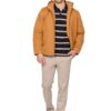 Man modeling a camel-colored jacket over a navy striped polo, beige chinos, and brown sneakers, standing with a hand in his pocket.