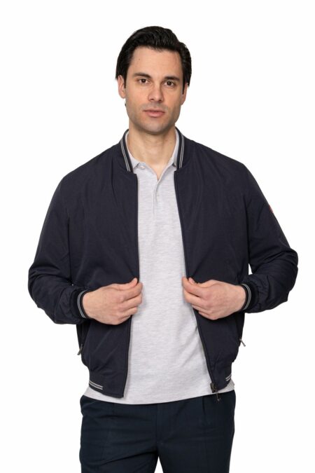 Man wearing a navy bomber jacket over a light grey polo, standing against a white background and holding the zipper.