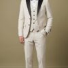 Man wearing a cream tuxedo with black satin lapels, bow tie, and pocket square, standing against a beige backdrop.