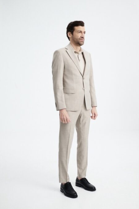Full-body shot of a man in a light beige suit and black dress shoes, standing and looking to the side in a studio setting.