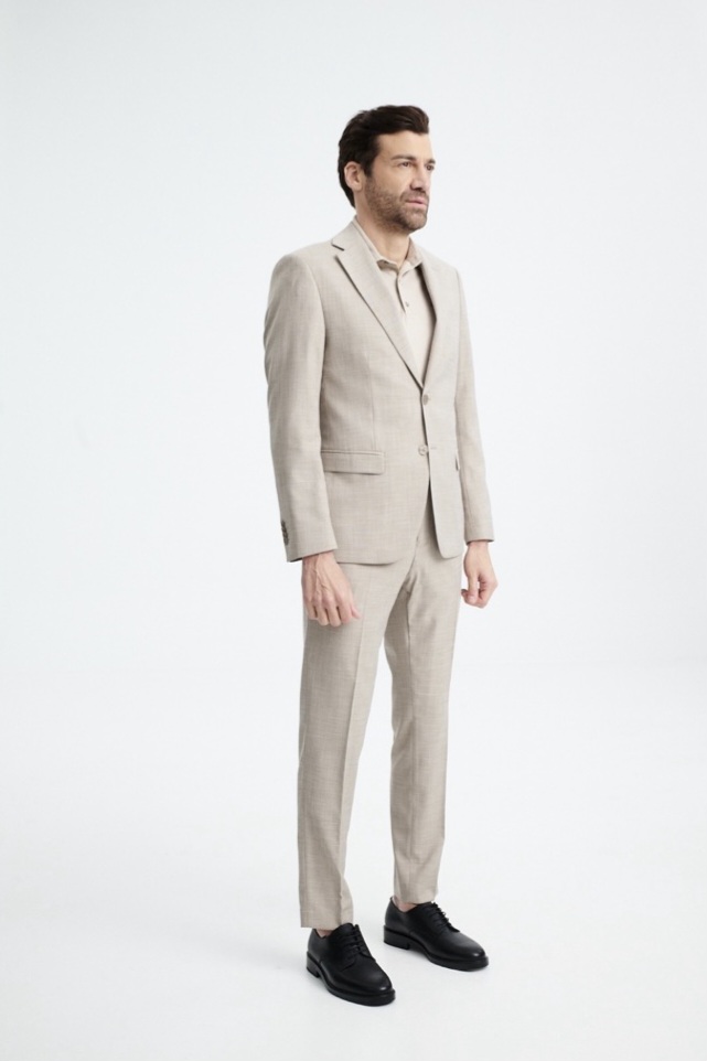 IMG-d15b84333595440d62be9572de9d94d5-V - Noble Full-body shot of a man in a light beige suit and black dress shoes, standing and looking to the side in a studio setting.
