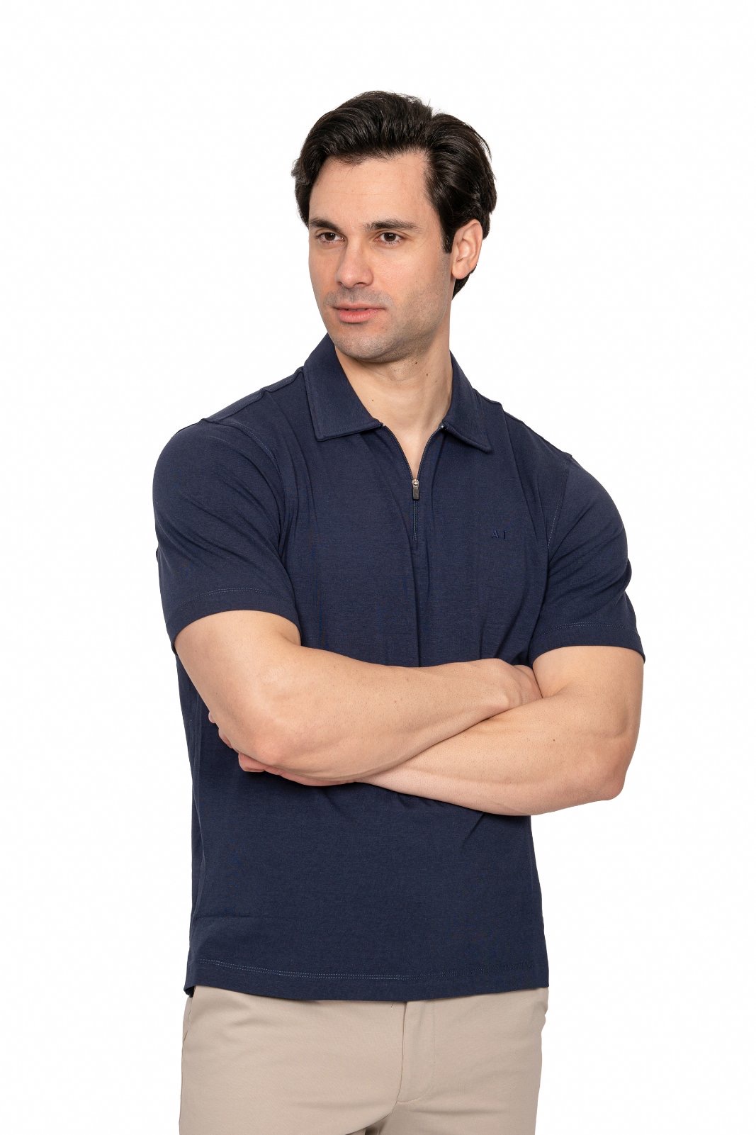 IMG_4921jpg - Noble Man with dark hair in a navy polo shirt and beige pants stands with arms crossed against a white background, looking to the side.