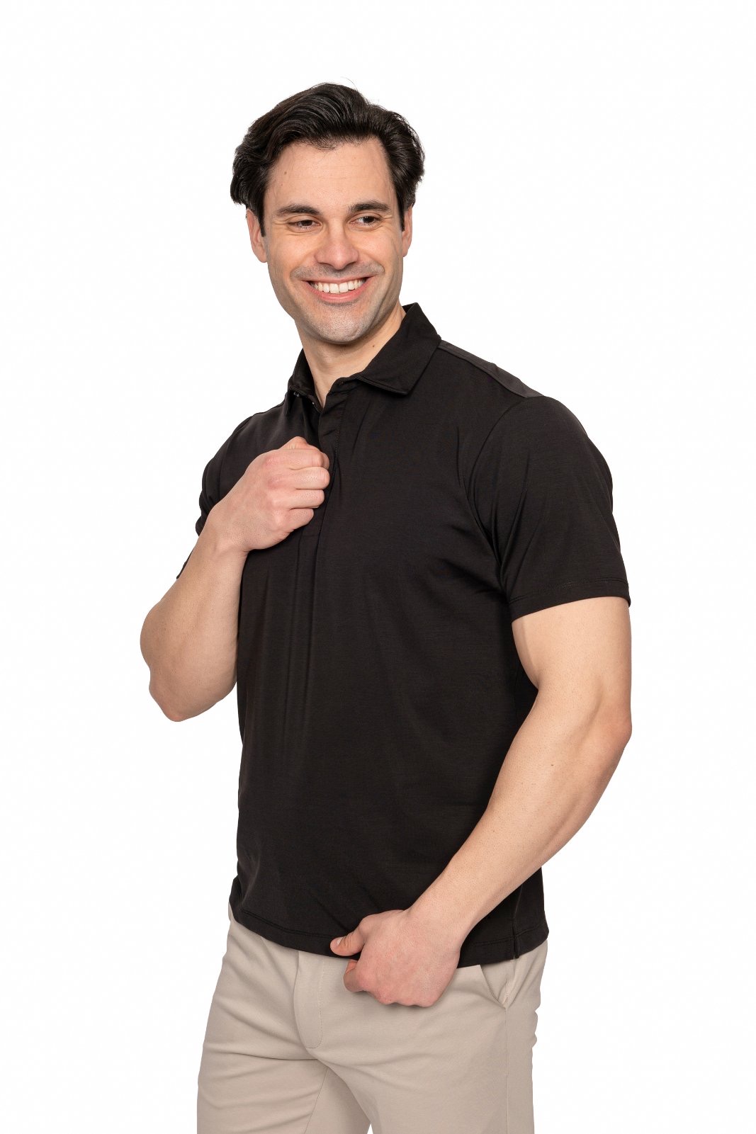 IMG_4977jpg - Noble Smiling man in a black polo shirt and beige pants, posing with his right hand on his chest against a white background.