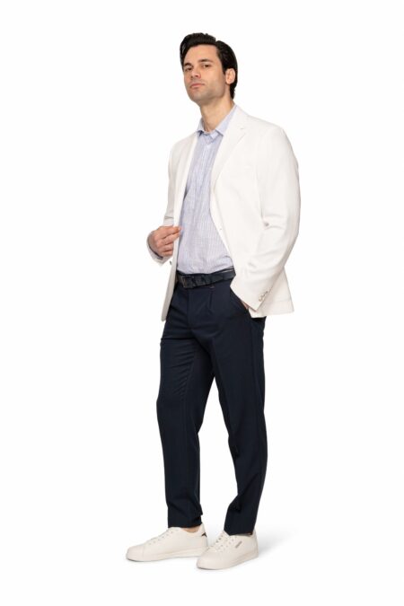 Man wearing a white blazer, light striped shirt, navy trousers, and white sneakers, posing with one hand on his jacket.